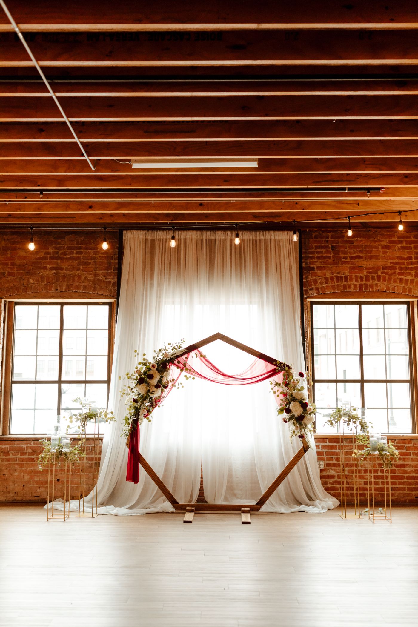 Industrial chic wedding ceremony setup featuring a geometric wooden arch adorned with red draping and floral arrangements, set against sheer white curtains and exposed brick walls with string lights at a loft-style event venue.