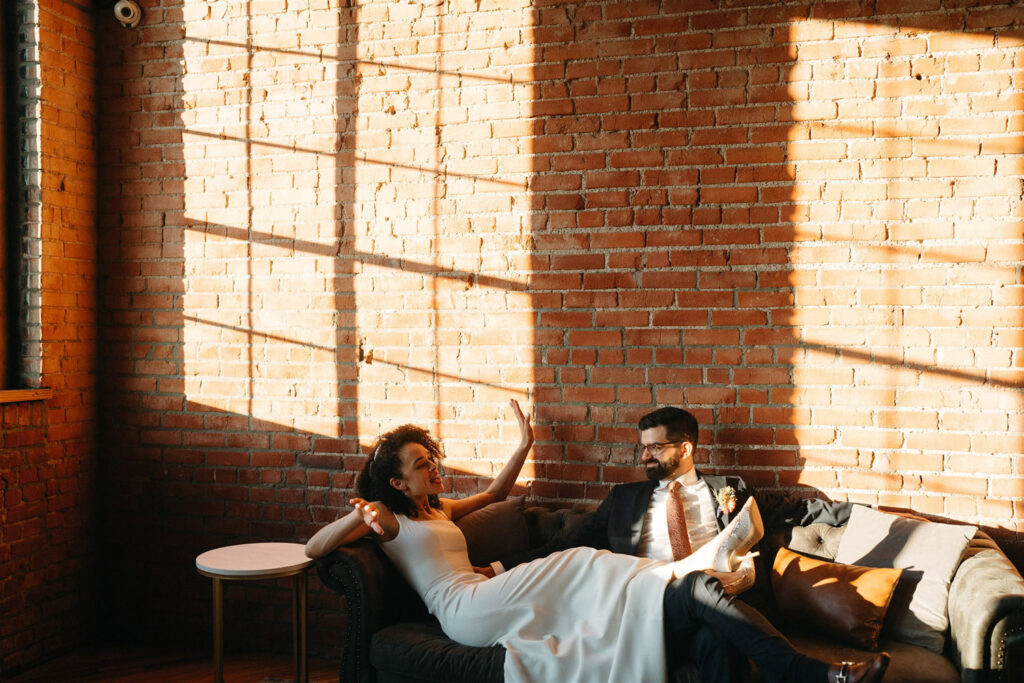 Bride and groom sitting next to each other on sofa with a brick wall as the background