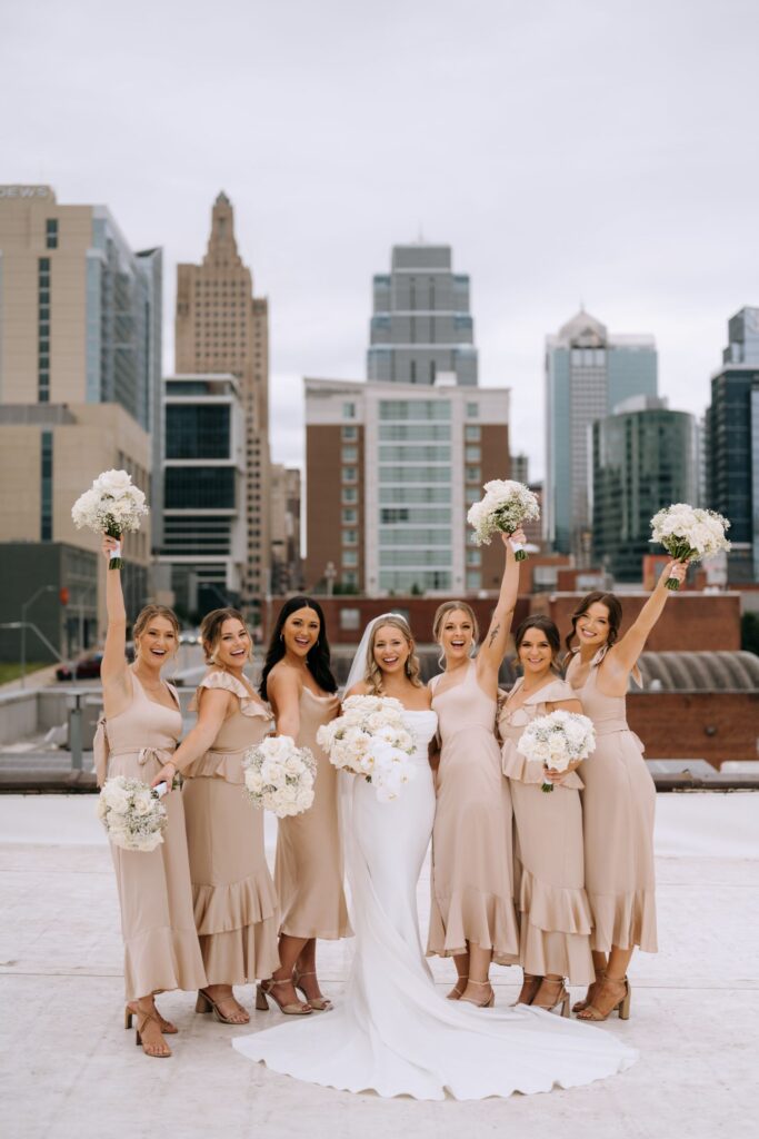 Bride and bridesmaids in champagne dresses posing on a Kansas City rooftop with the city skyline in the background.