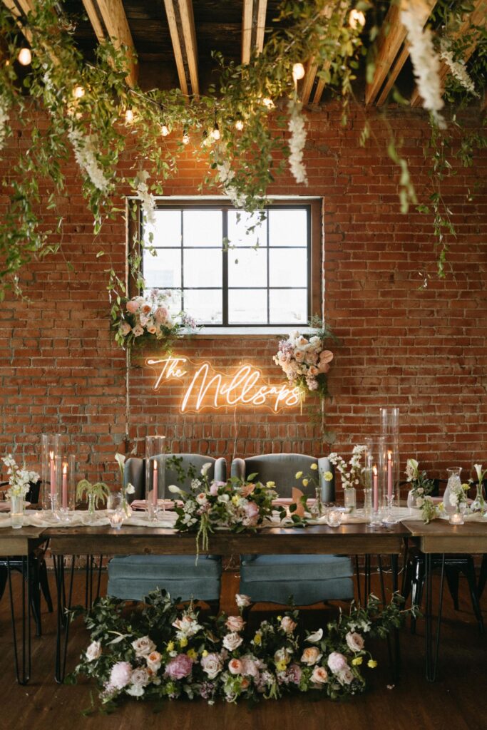 Detail of a wedding reception table at UNION in Kansas City, MO, with floral arrangements, candles, and a custom neon sign on a brick wall.