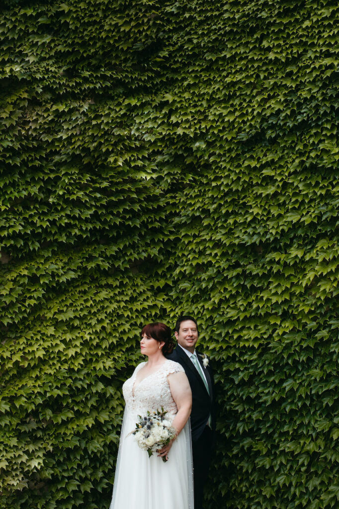 A bride and groom posing for wedding day photos against a greenery wall.