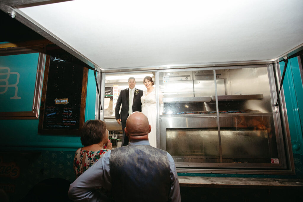 A bride and groom standing inside a mobile ice cream truck.