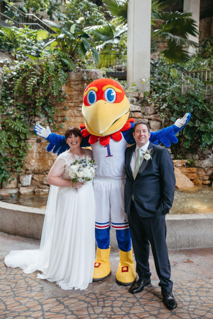 A bride and groom on their wedding day posing for a photo with the groom's college mascot, Big Jay from the University of Kansas.
