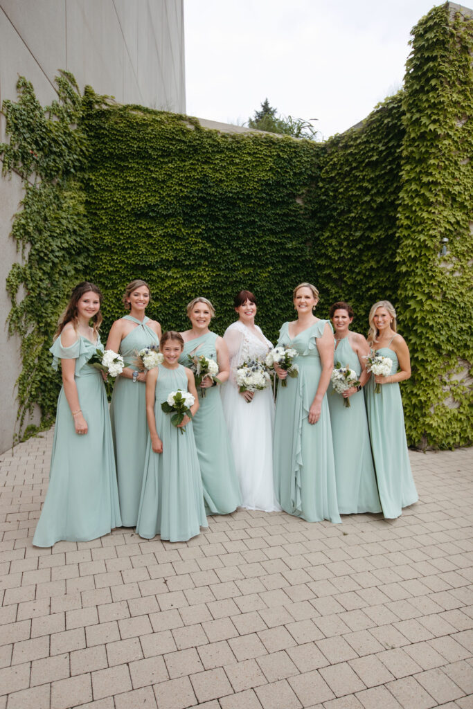 A bride with her bridesmaids wearing pale green dresses surrounded by a wall of greenery.